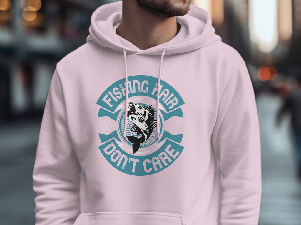 In an urban setting, someone sports a Garment Graphics light pink hoodie with the phrase Fishing Hair Dont Care and an anglers catch at its center. The blurred background highlights city buildings.