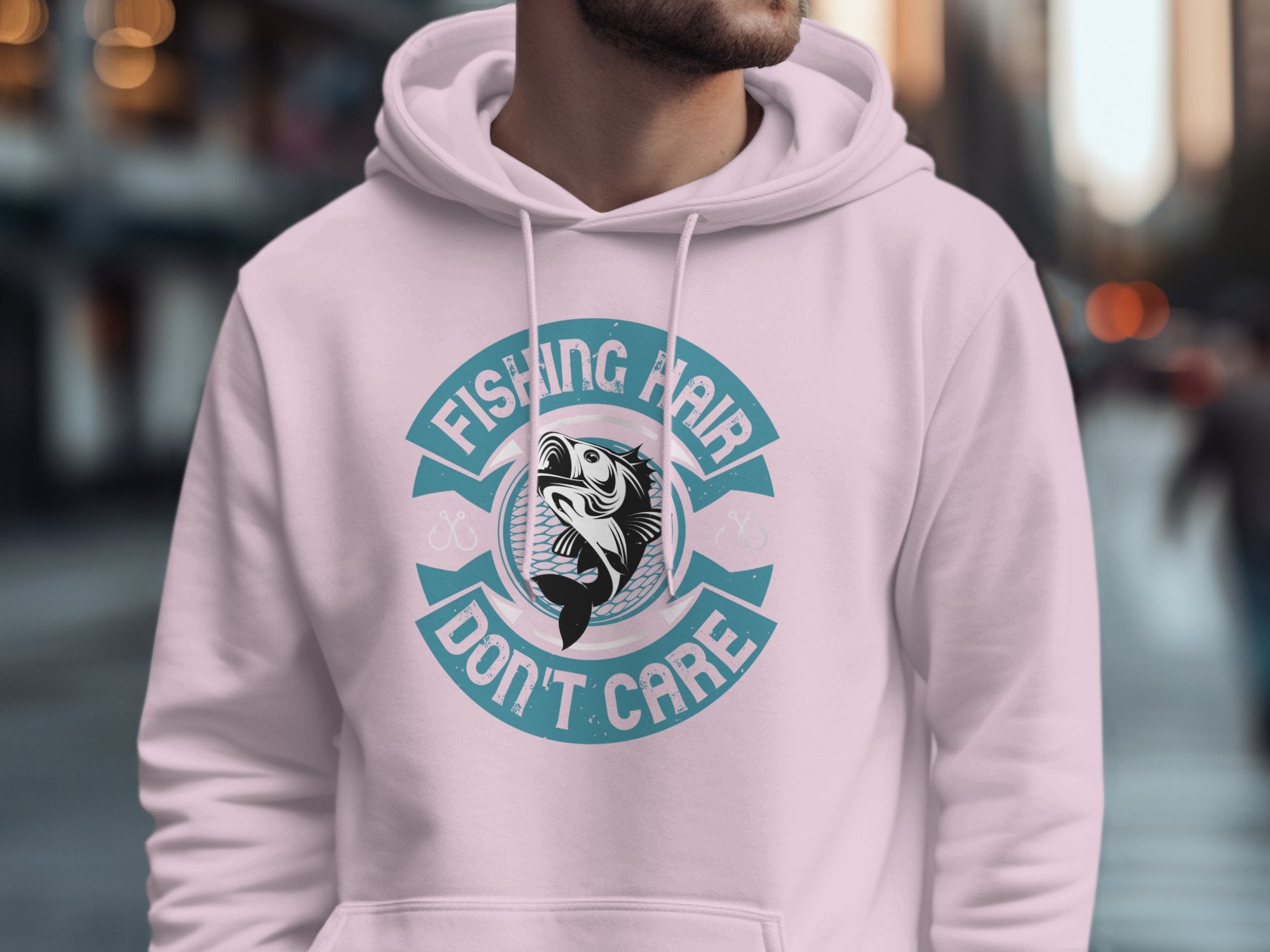 In an urban setting, someone sports a Garment Graphics light pink hoodie with the phrase Fishing Hair Dont Care and an anglers catch at its center. The blurred background highlights city buildings.