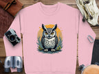 This pink unisex sweatshirt from Garment Graphics features an owl with an intense gaze against an orange and yellow circle. Styled with a camera, notebook, binoculars, bag, and climbing gear on a wooden surface.
