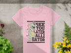 Garment Graphics presents the Alligator T-Shirt for women, featuring a whimsical cartoon of a smiling alligator with a basketball and the caption See you later, alligator, against a concrete backdrop with green plants and yellow flowers—ideal for humor lovers.