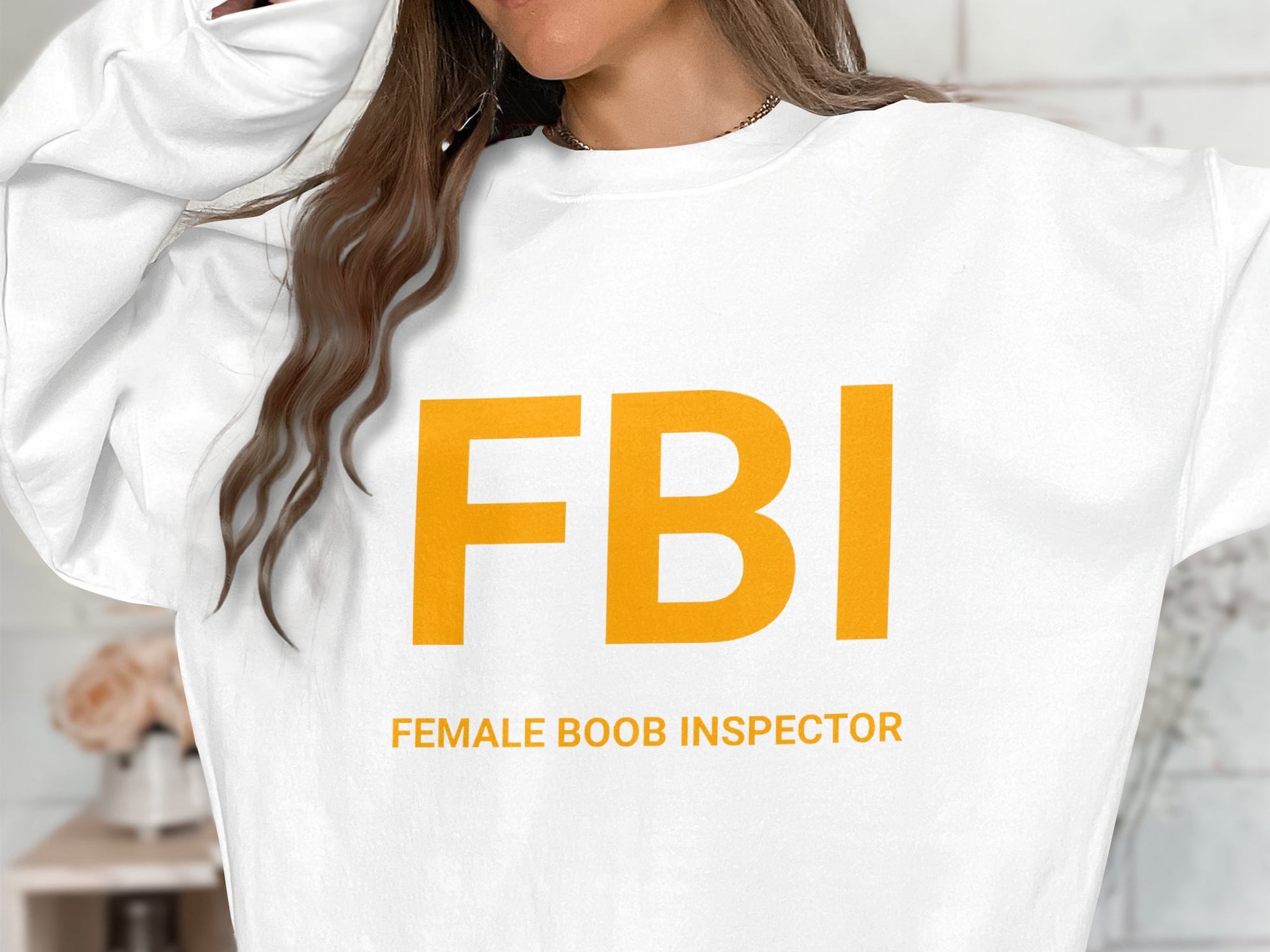 A long-haired individual raises their hand to the side of their head, wearing a Garment Graphics sweatshirt with large yellow FBI lettering that humorously reads Female Boob Inspector, available in unisex sizes for a perfect fit.