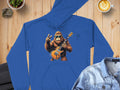 The Garment Graphics blue music lovers hoodie, featuring a cartoon gorilla with an electric guitar and peace sign, is stylishly displayed on a wooden surface beside a denim jacket, latte art, and leafy plants.