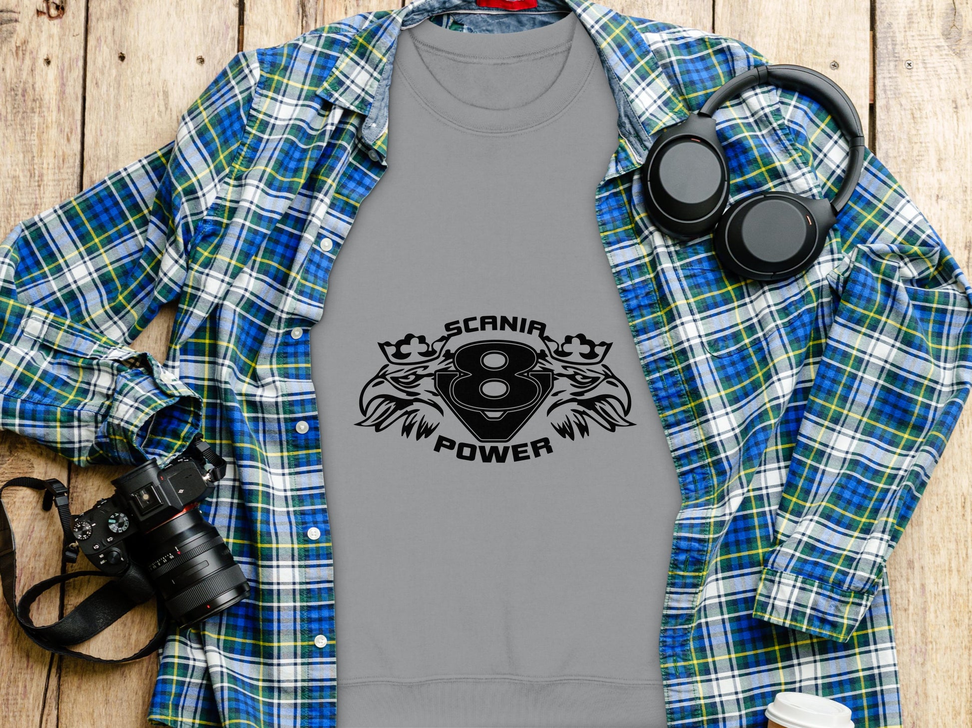 A Garment Graphics gray t-shirt with the Scania Power 8 logo and twin eagle designs is placed on a plaid shirt. Surrounding the set are headphones, a camera, and a white to-go cup, all neatly arranged on a wooden surface.