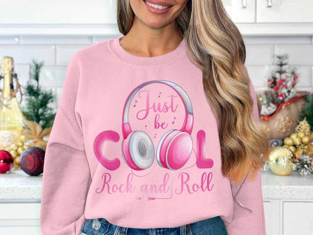 A woman in a kitchen wears a pink sweatshirt by Garment Graphics, featuring Rock and Roll Headphones and musical notes. The text says, Just be cool, rock and roll. Holiday decor like ornaments and pine branches set a festive scene for music enthusiasts or fashion lovers.