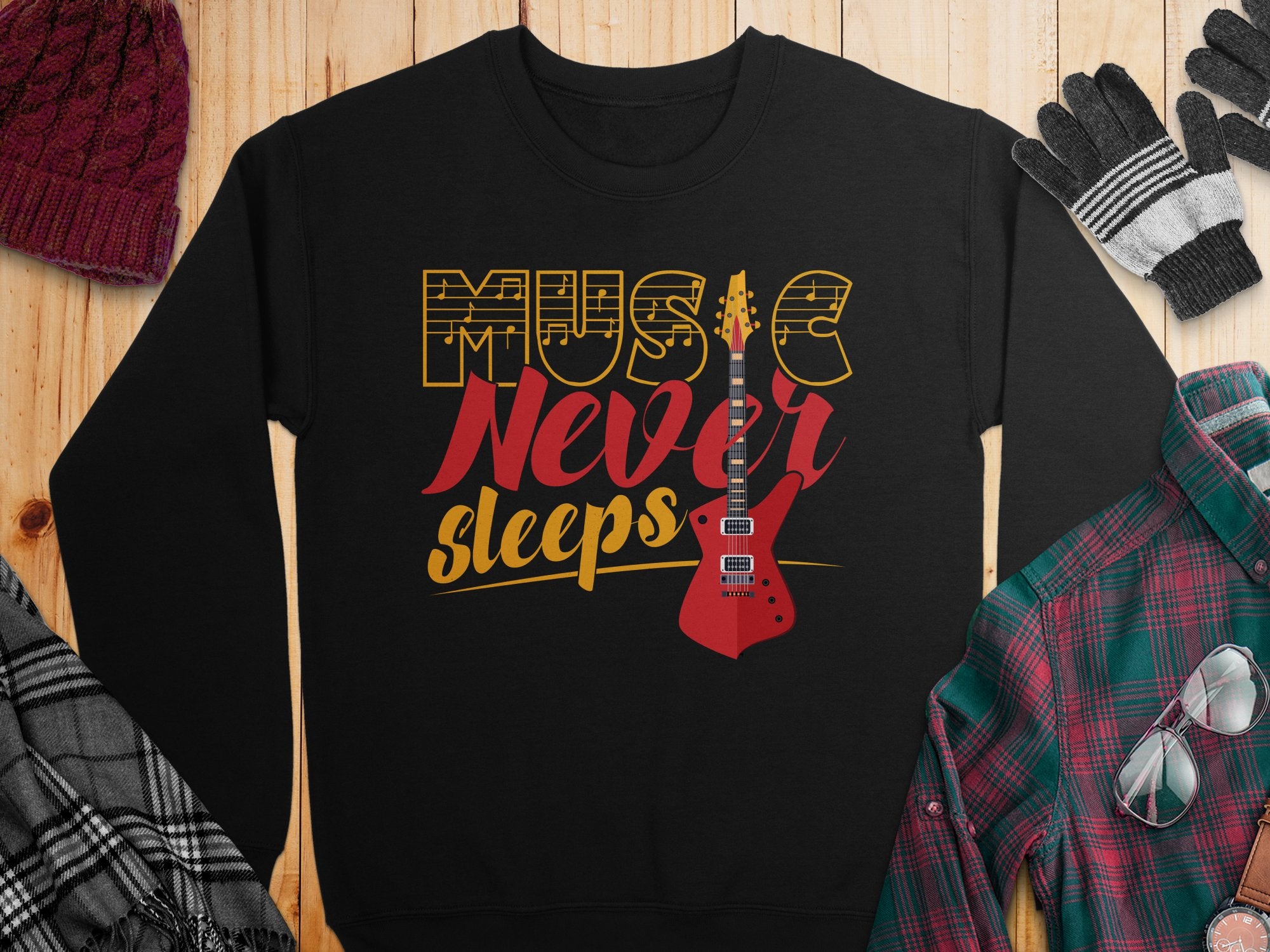 The Garment Graphics sweatshirt features Music Never Sleeps Guitar in bold yellow and red with a red electric guitar graphic. Styled on wood with a knit hat, gloves, plaid shirt, and scarf, it’s perfect for music lovers.