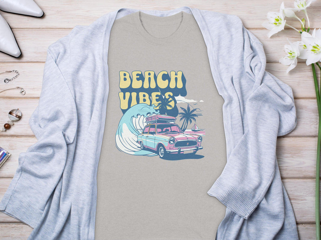 Garment Graphics gray graphic t-shirt with BEACH VIBES text, classic car, surfboards, waves, and palm trees design. Its on a light wooden surface under a light gray cardigan. Nearby are flowers, jewelry, and shoes.