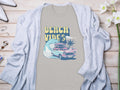 Garment Graphics gray graphic t-shirt with BEACH VIBES text, classic car, surfboards, waves, and palm trees design. Its on a light wooden surface under a light gray cardigan. Nearby are flowers, jewelry, and shoes.