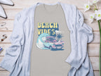 Garment Graphics gray graphic t-shirt with BEACH VIBES text, classic car, surfboards, waves, and palm trees design. Its on a light wooden surface under a light gray cardigan. Nearby are flowers, jewelry, and shoes.