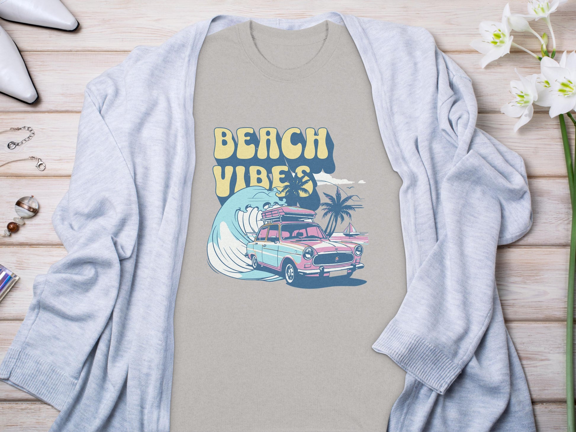 Garment Graphics gray graphic t-shirt with BEACH VIBES text, classic car, surfboards, waves, and palm trees design. Its on a light wooden surface under a light gray cardigan. Nearby are flowers, jewelry, and shoes.