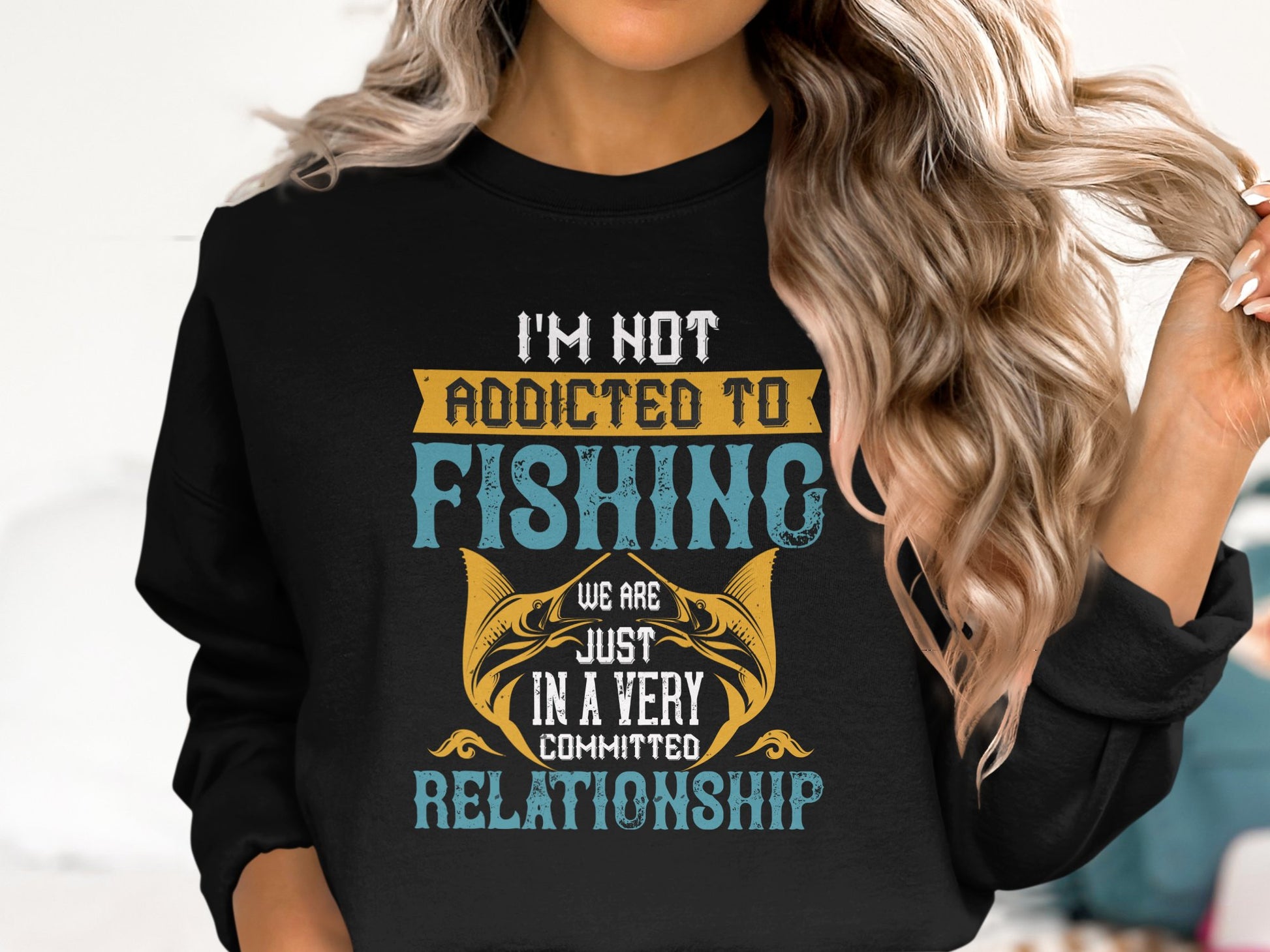 A woman with long, wavy blonde hair wears a Garment Graphics black sweatshirt in unisex sizing. It showcases yellow and blue text that says, Im not addicted to fishing, we are just in a very committed relationship, alongside a fish design.