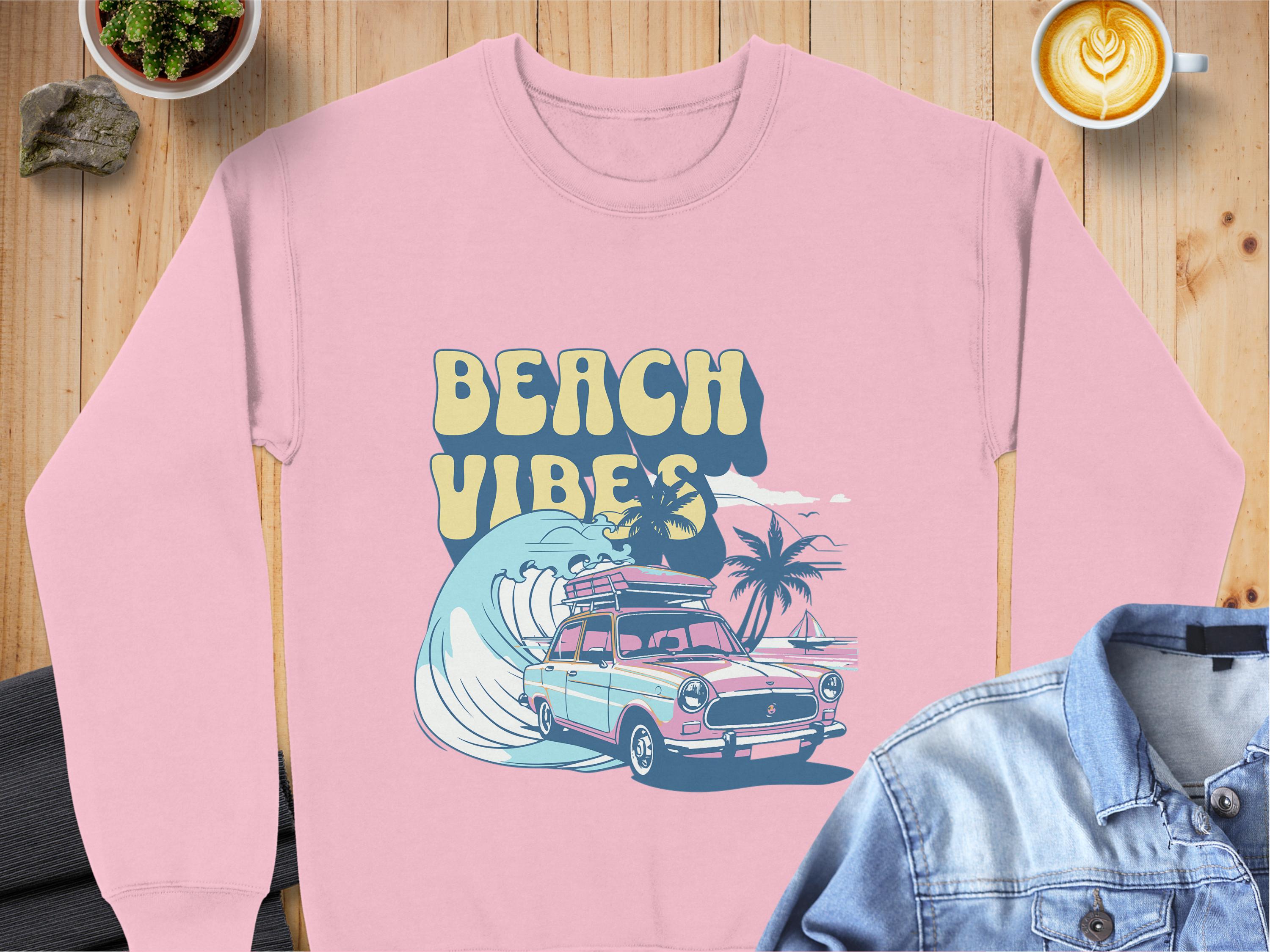 A pink graphic sweatshirt featuring a retro car, surfboards, waves, and palm trees rests on the table. Ideal for a road trip, it displays BEACH VIBES in unisex sizing by Garment Graphics. A denim jacket, heart-design latte, and potted plant are nearby.