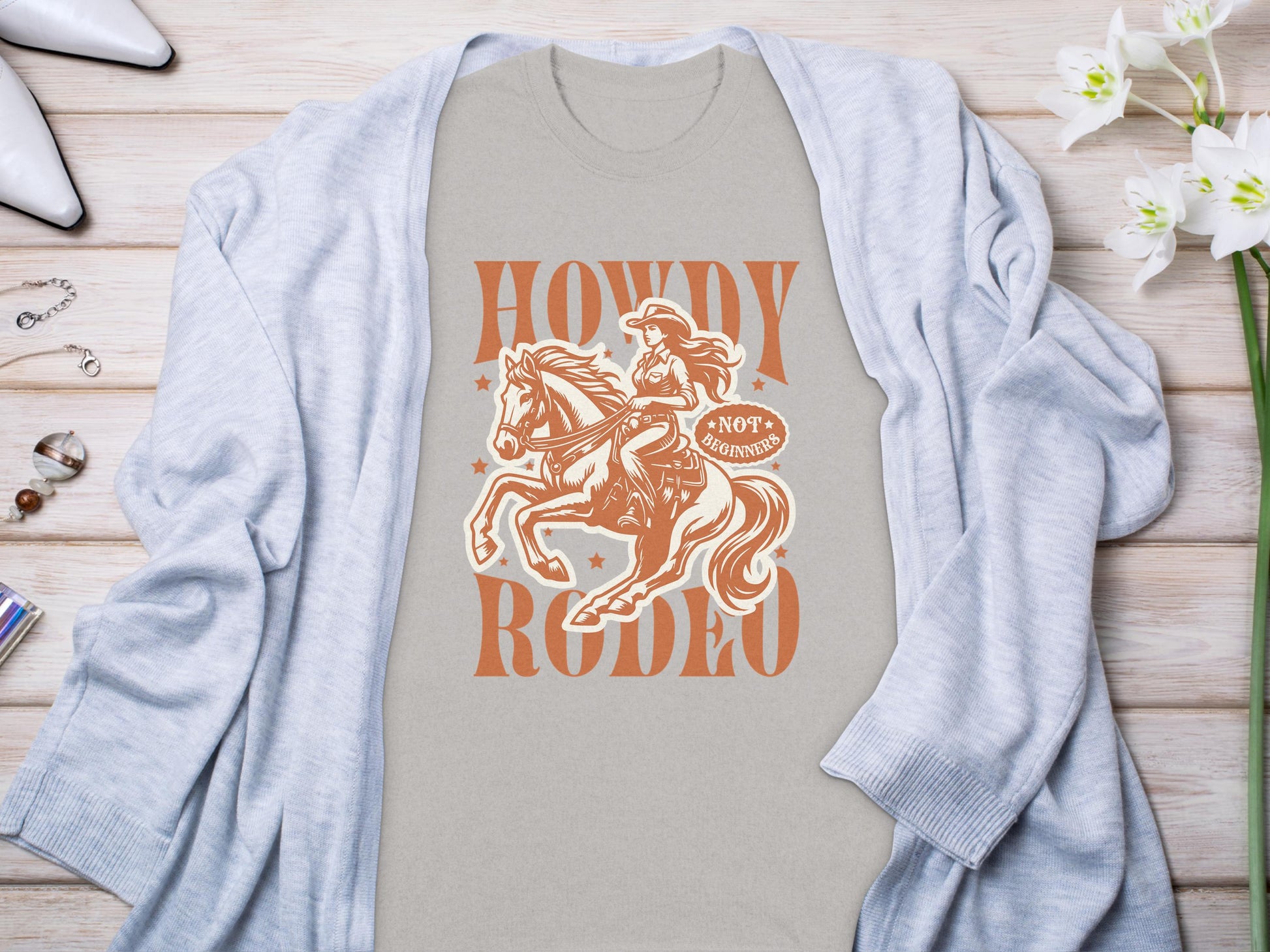 A light gray Garment Graphics Howdy Rodeo T-Shirt with a cowboy on a horse graphic lies beneath a pastel blue cardigan on wood. White high heels, an elegant bracelet, rings, and delicate white flowers complete this ensemble for enthusiasts.