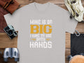 A Garment Graphics gray classic fit tee lies on a wooden surface, featuring the humorous message Mine is so big I have to use both hands in bold white and yellow text. Surrounding it are a camera, binoculars, leather pouch, watch, and map.