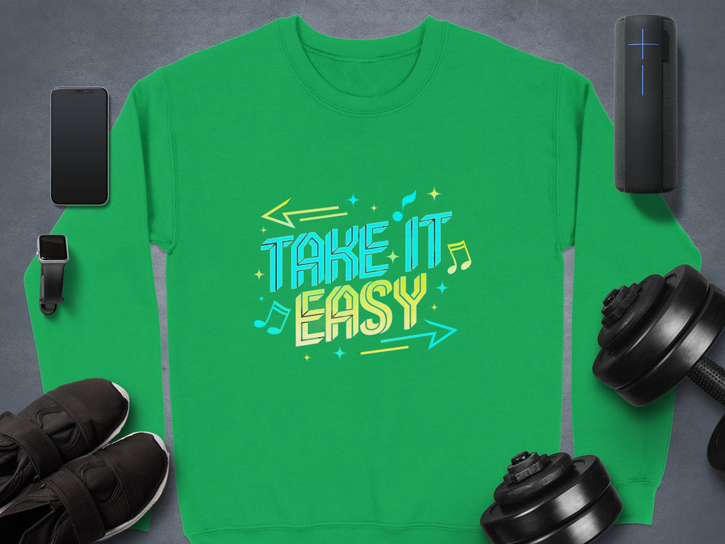 Unisex comfy sweatshirt in green by Garment Graphics features TAKE IT EASY with musical notes and arrows, ideal for casual wear. Displayed on gray with a smartphone, smartwatch, portable speaker, black sneakers, and dumbbells.