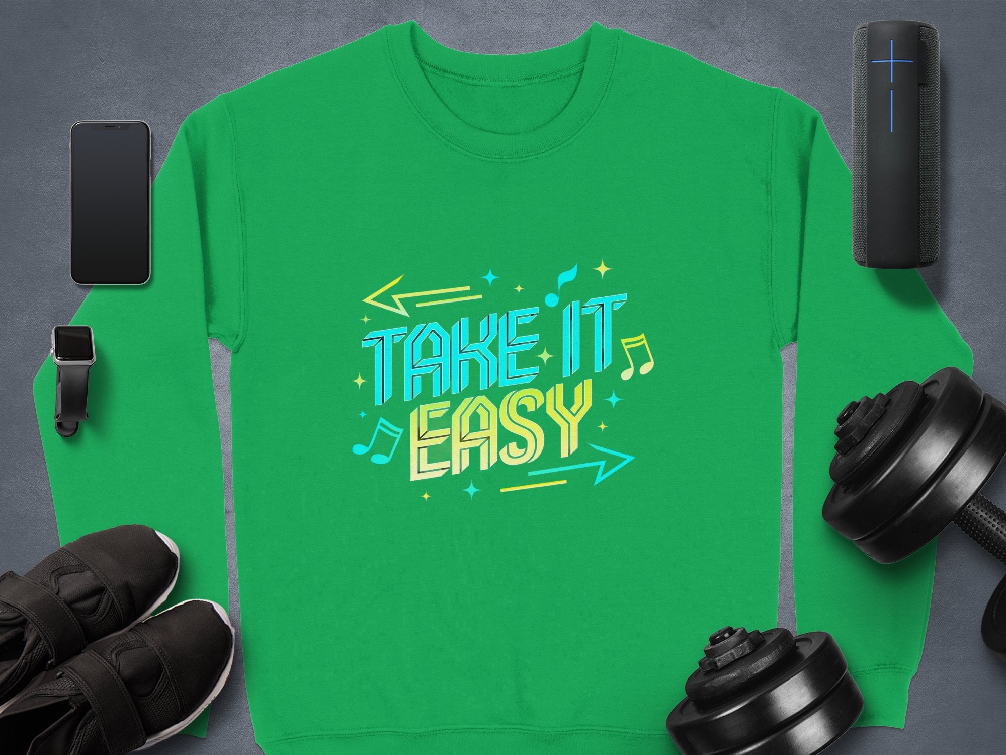 Unisex comfy sweatshirt in green by Garment Graphics features TAKE IT EASY with musical notes and arrows, ideal for casual wear. Displayed on gray with a smartphone, smartwatch, portable speaker, black sneakers, and dumbbells.