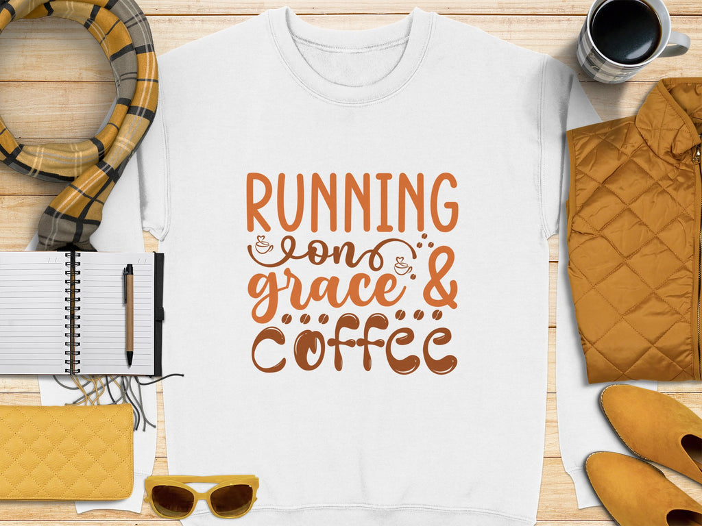 A Garment Graphics white t-shirt featuring Running on Grace & Coffee in bold orange lies on wood, surrounded by a notebook, pen, plaid scarf, mug, quilted jacket, yellow wallet, and sunglasses. Perfect unisex sizing for those fueled by grace and coffee!.