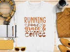 A Garment Graphics white t-shirt featuring Running on Grace & Coffee in bold orange lies on wood, surrounded by a notebook, pen, plaid scarf, mug, quilted jacket, yellow wallet, and sunglasses. Perfect unisex sizing for those fueled by grace and coffee!.