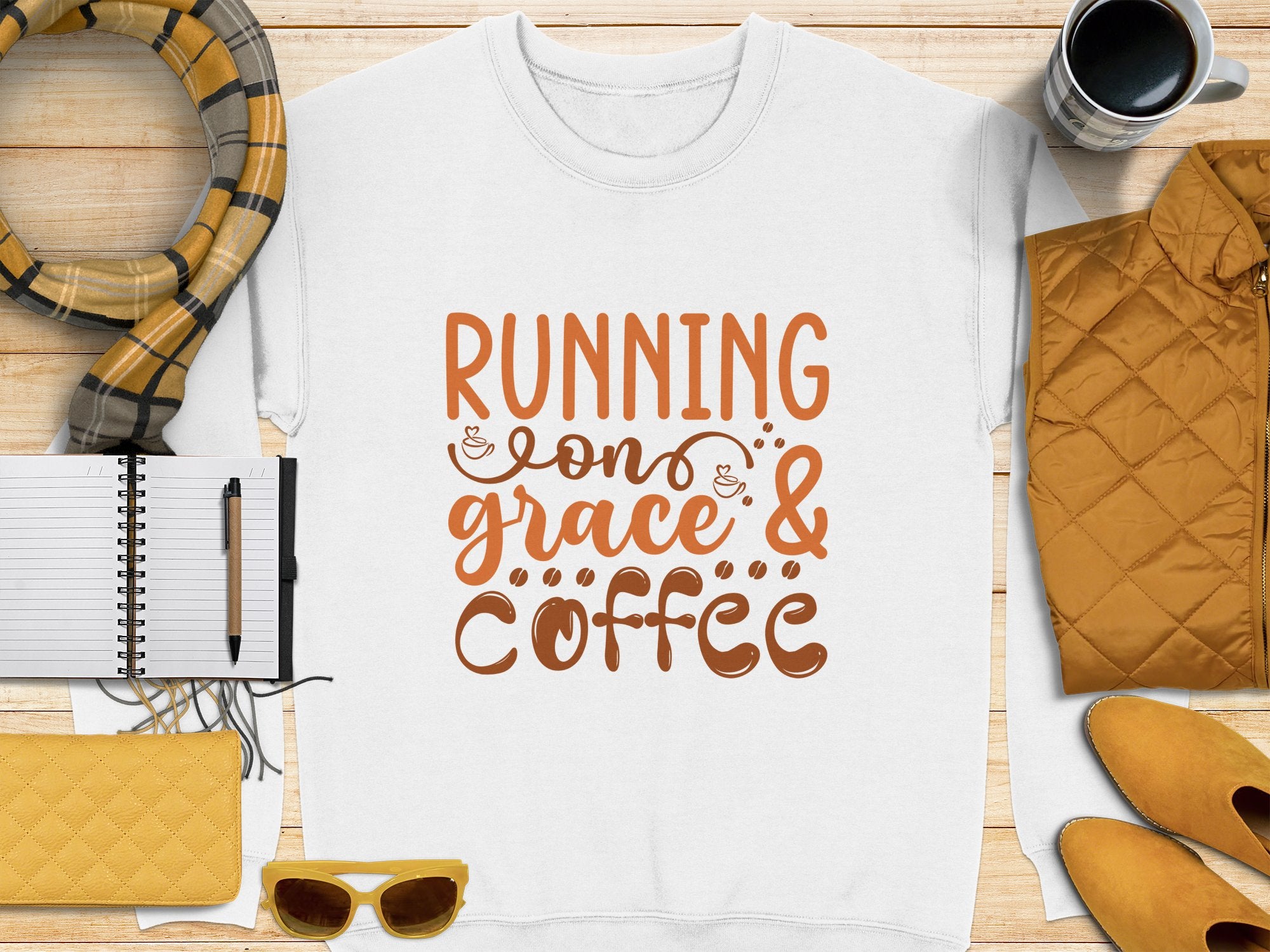 A Garment Graphics white t-shirt featuring Running on Grace & Coffee in bold orange lies on wood, surrounded by a notebook, pen, plaid scarf, mug, quilted jacket, yellow wallet, and sunglasses. Perfect unisex sizing for those fueled by grace and coffee!.