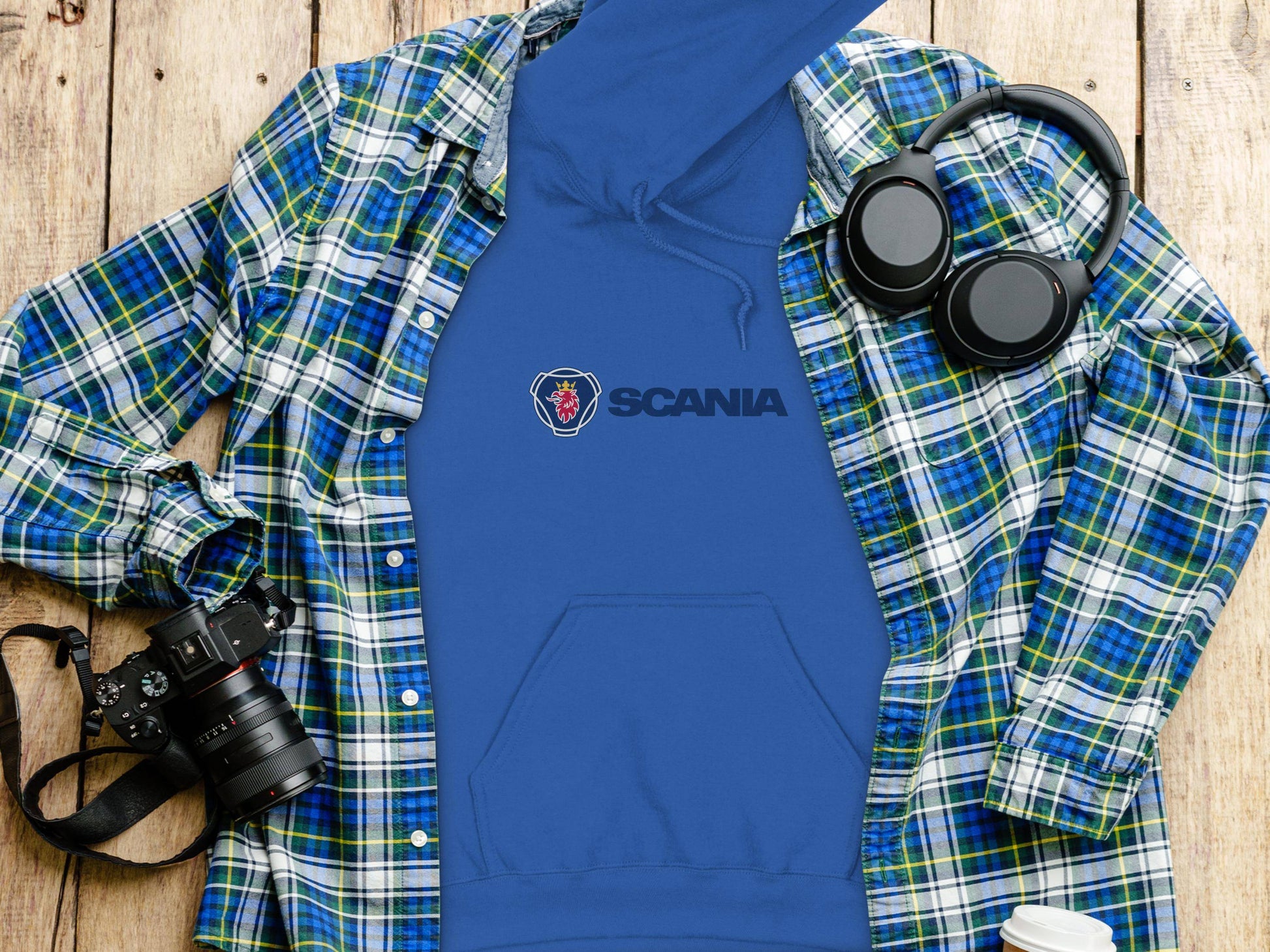 A blue Scania logo hoodie from Garment Graphics lies on a wooden surface, elegantly draped with a green and blue plaid shirt. Nearby, black headphones, a camera, and a white travel mug are artfully arranged for the perfect flat lay photo.