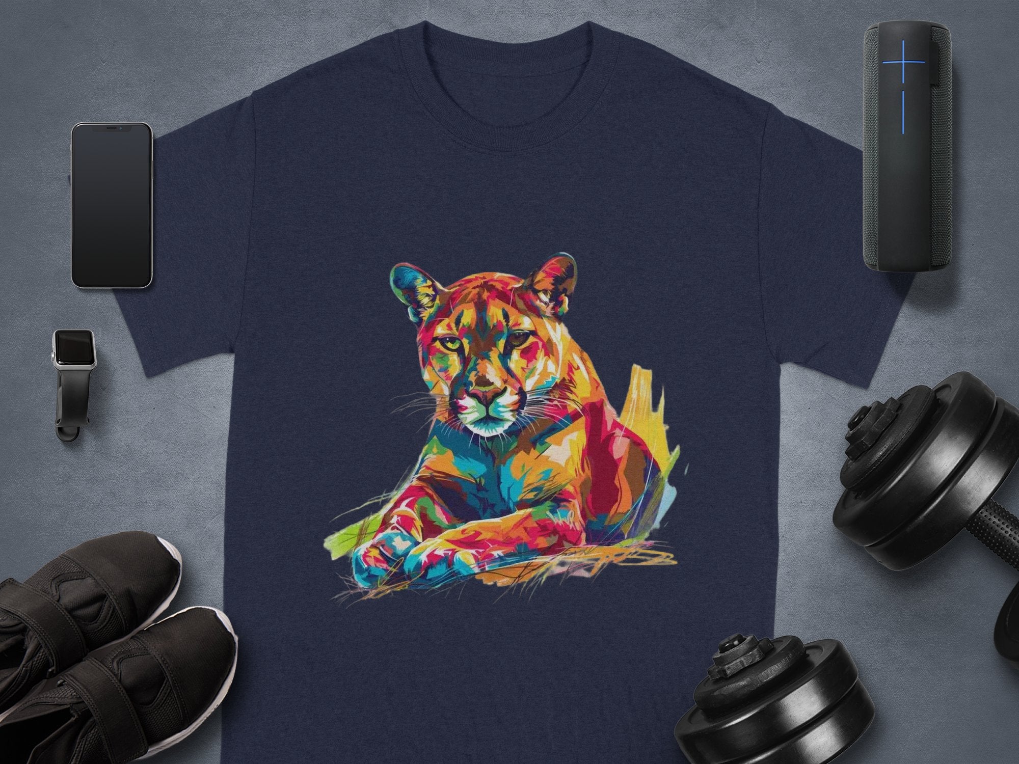 A dark blue T-shirt by Garment Graphics showcases a vibrant roaring cougar, surrounded by a smartphone, smartwatch, black sneakers, wireless speaker, and adjustable dumbbells on a gray backdrop.
