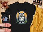 A black unisex sweatshirt by Garment Graphics, featuring an owl design in the center, is displayed on a wooden background surrounded by accessories: sunglasses, makeup brushes, eyeshadow palette, red lipstick, nail polish, pink shoes, a blue pouch, and a yellow blazer.