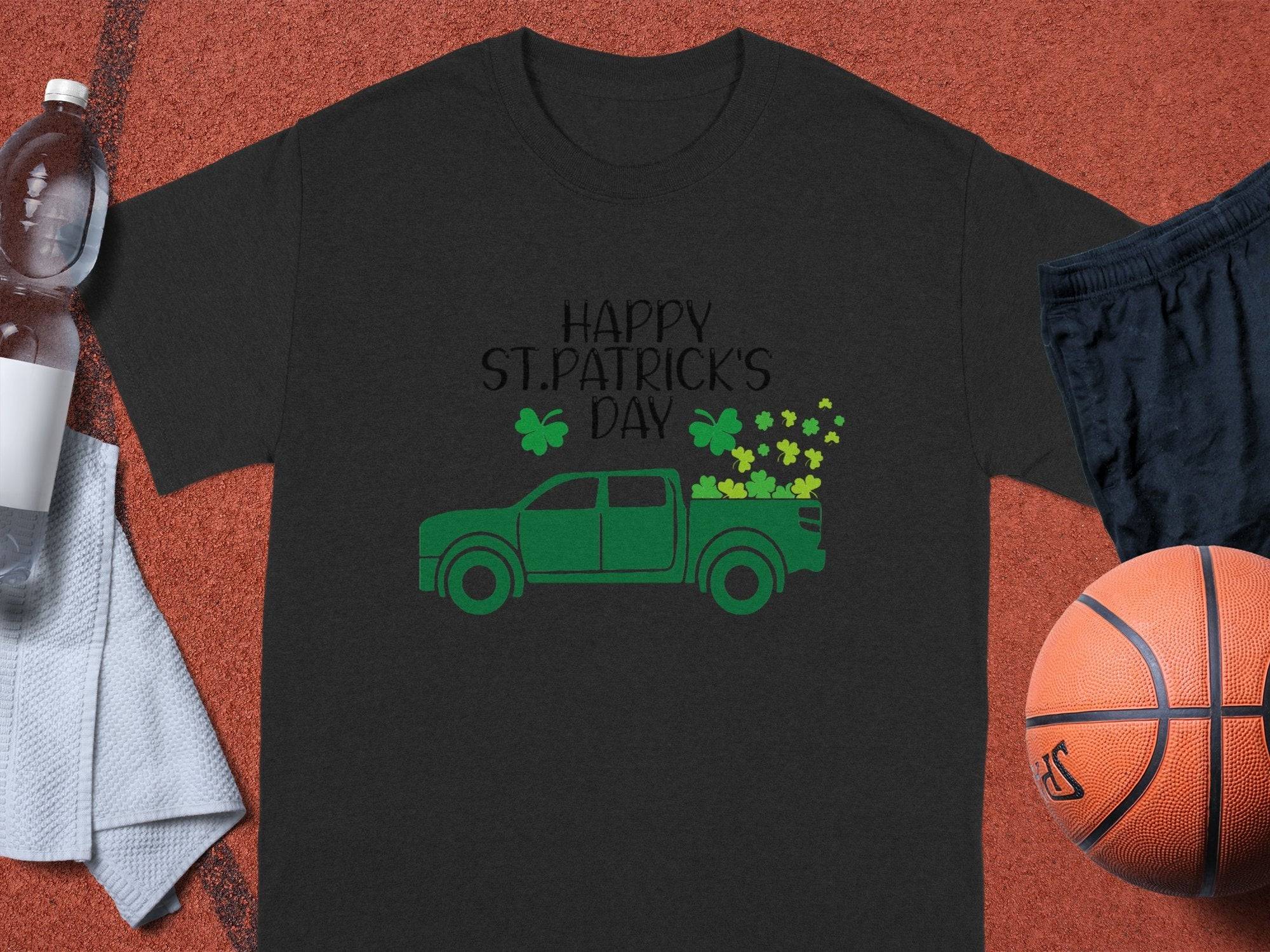 A Garment Graphics black T-shirt celebrates Irish pride with a green pickup and shamrocks. Happy St. Patricks Day is printed above, while a basketball, black shorts, clear water bottle, and white towel are in the background on a sports court.