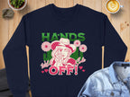 Garment Graphics presents a navy blue, loose-fit sweatshirt featuring a person in a hat with a finger on their lips, adorned with bold Hands Off Wild Side Sweatshirts text surrounded by flowers and hearts. Laid on wood, it pairs perfectly with a denim jacket and latte.
