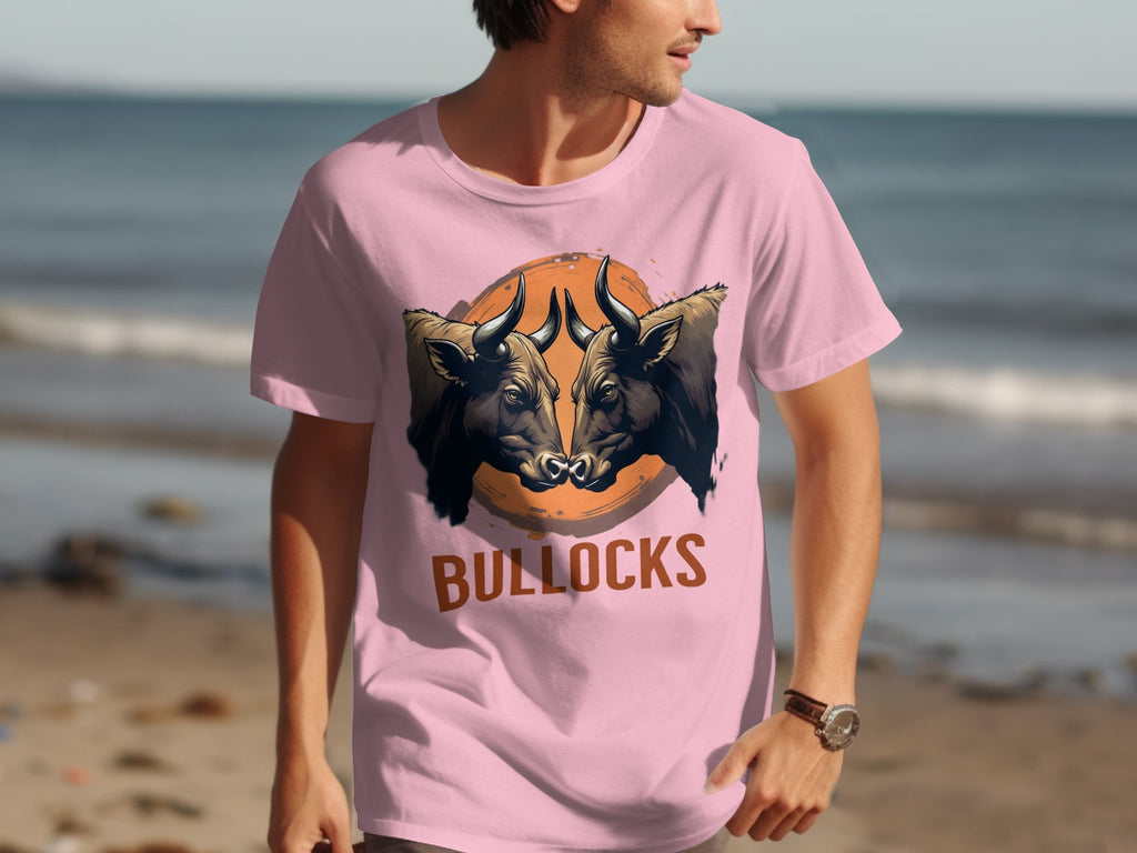 A man in a classic fit pink T-shirt by Garment Graphics, featuring two bulls and BULLOCKS in bold lettering, walks along the sandy beach with the ocean behind him.