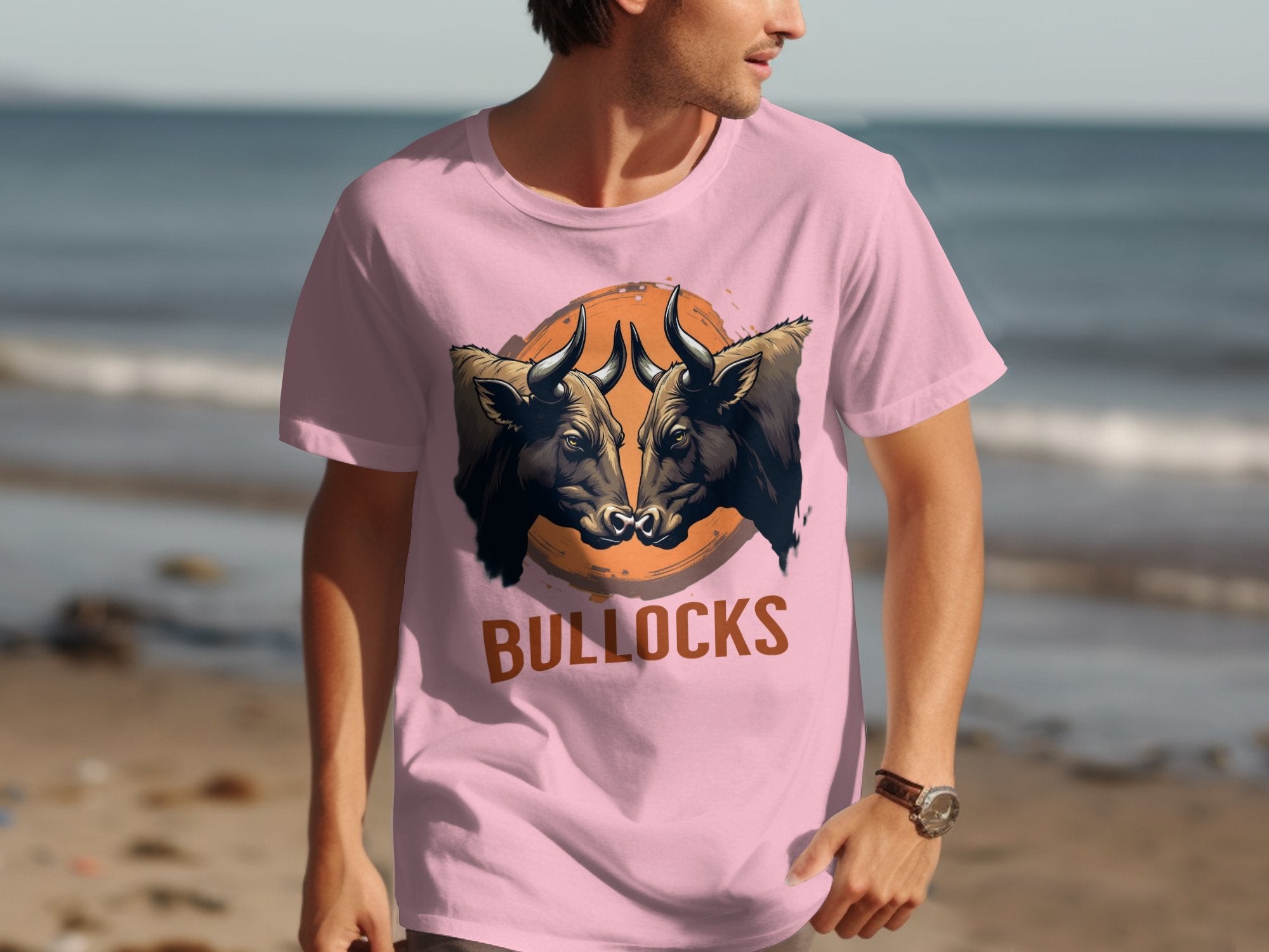 A man in a classic fit pink T-shirt by Garment Graphics, featuring two bulls and BULLOCKS in bold lettering, walks along the sandy beach with the ocean behind him.