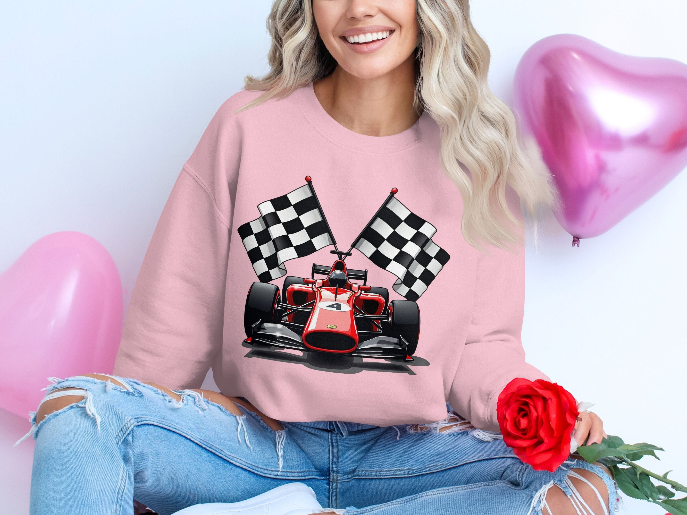 A woman in a vibrant pink Garment Graphics sweatshirt featuring a red racing car and checkered flags sits cross-legged. With long blonde hair and holding a red rose, she poses in front of two pink heart-shaped balloons—ideal for motorsport enthusiasts.