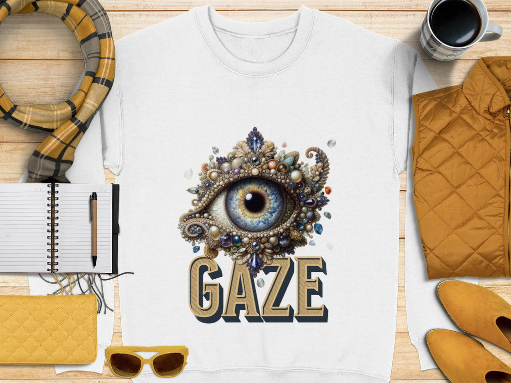 Garment Graphics offers a white unisex t-shirt with an ornate eye design featuring a blue eye surrounded by jewels above GAZE. Accompanied by a notebook, pen, mug, quilted jacket, scarf, yellow sunglasses, and matching wallet on a wooden surface.