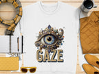 Garment Graphics offers a white unisex t-shirt with an ornate eye design featuring a blue eye surrounded by jewels above GAZE. Accompanied by a notebook, pen, mug, quilted jacket, scarf, yellow sunglasses, and matching wallet on a wooden surface.