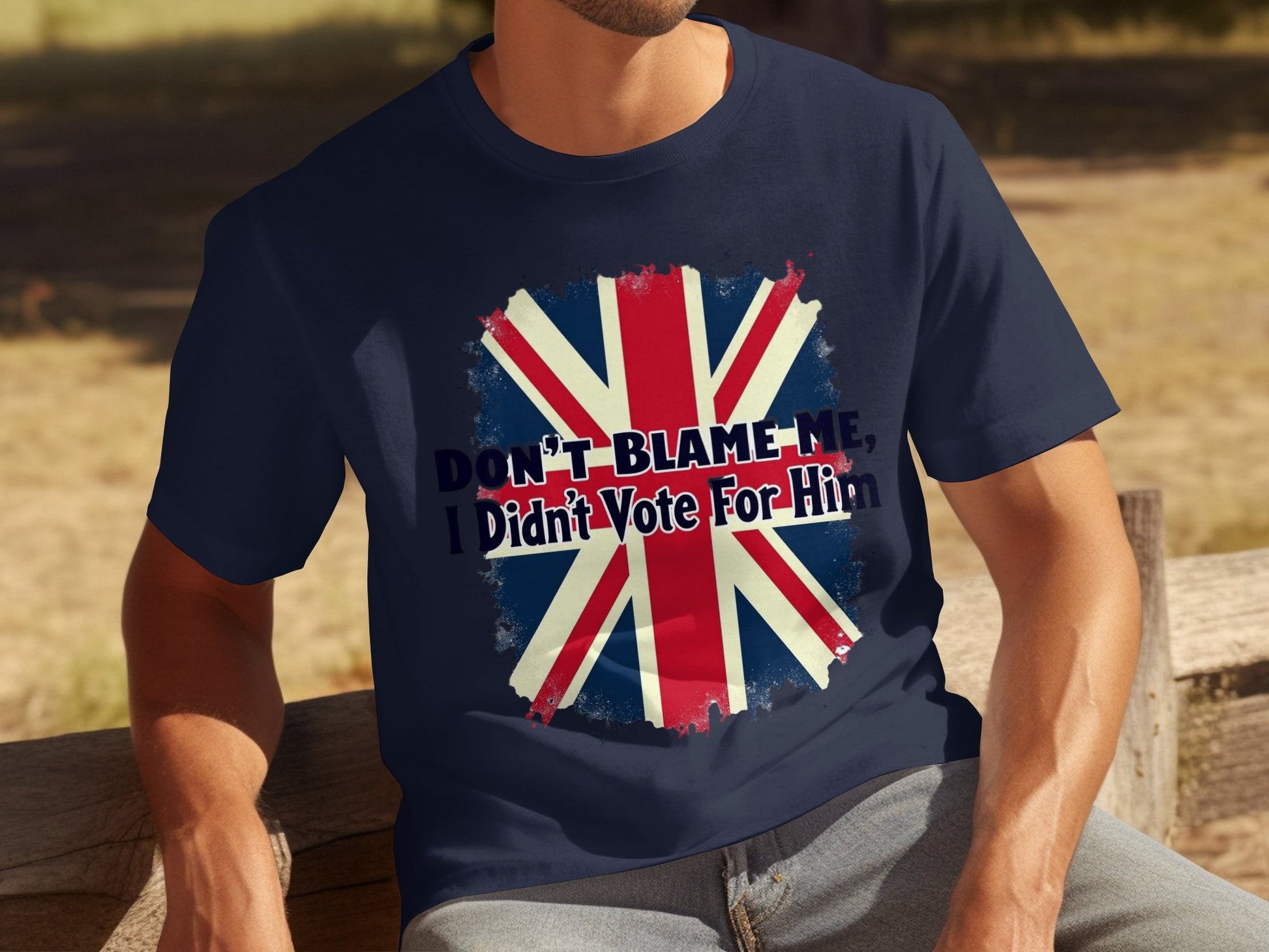 A person sits on a bench in a classic fit navy blue T-shirt by Garment Graphics, showcasing the Union Jack with the bold statement, Dont blame me, I didnt vote for him, above it. The individuals face is not visible in the image.