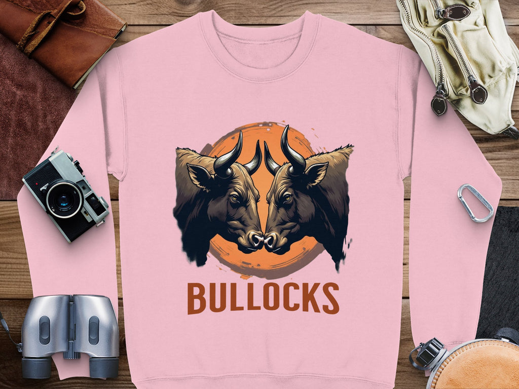 The loose-fit sweatshirt from Garment Graphics features a striking bullocks graphic with two bulls facing each other over an orange circle and BULLOCKS text, displayed on a wooden surface alongside binoculars, a camera, and travel items.