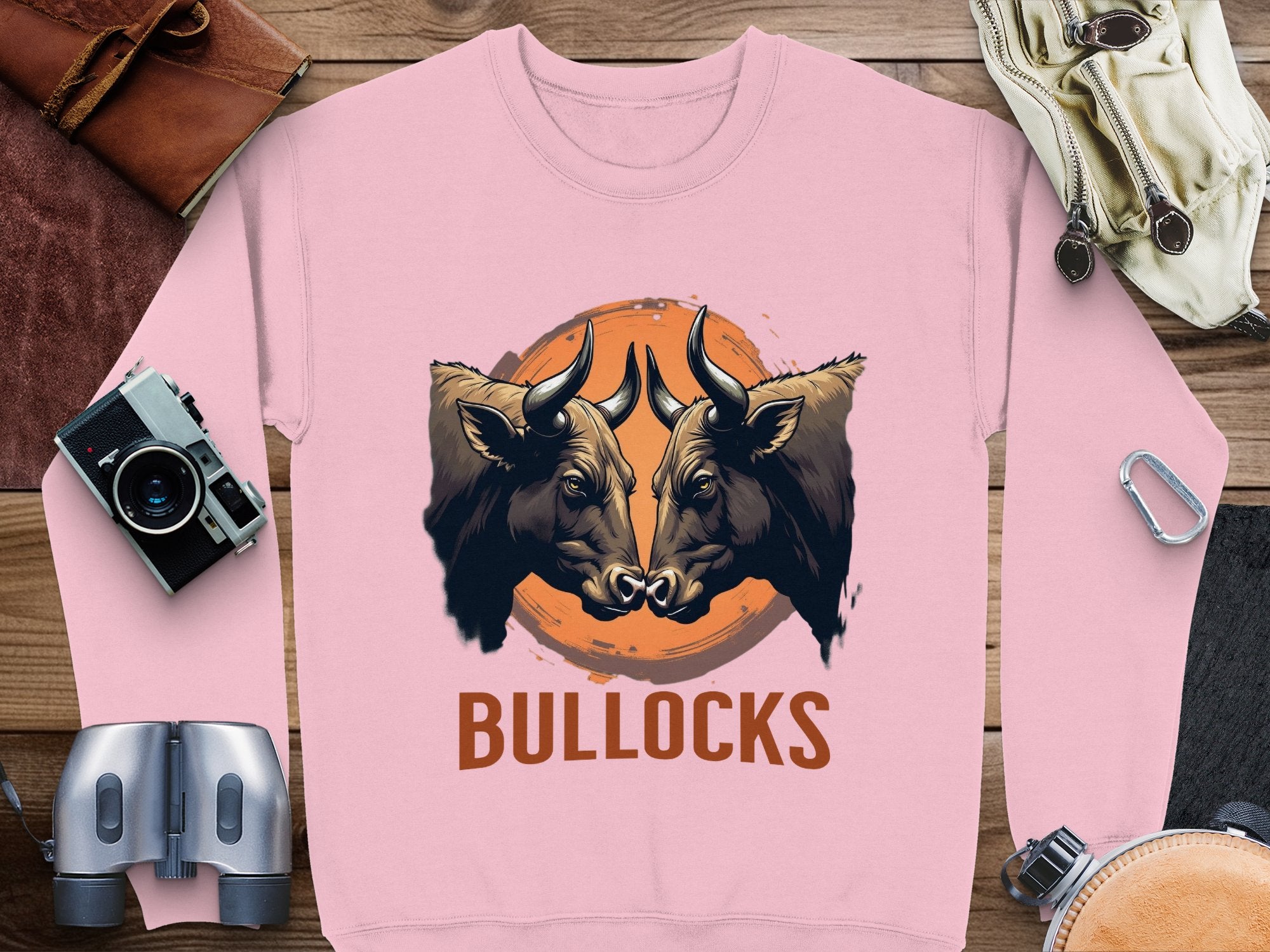 The loose-fit sweatshirt from Garment Graphics features a striking bullocks graphic with two bulls facing each other over an orange circle and BULLOCKS text, displayed on a wooden surface alongside binoculars, a camera, and travel items.