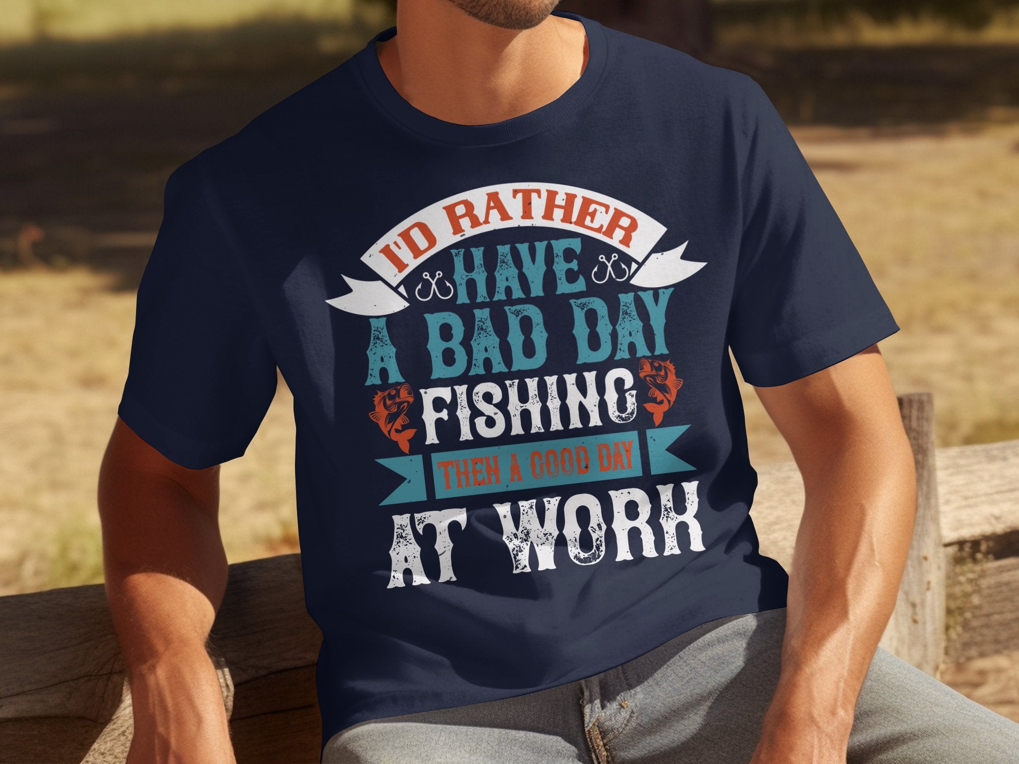 A person on a wooden bench wears a Garment Graphics fishing T-shirt with the message, Id rather have a bad day fishing than a good day at work, featuring decorative blue, white, and orange text perfect for anglers.
