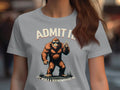 A person wears a light gray T-shirt from Garment Graphics with a cartoon Bigfoot design. It reads ADMIT IT BIGFOOT on top and LIFE WOULD BE BORING WITHOUT ME below, set against a blurred background that emphasizes the lively graphic.