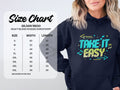 A woman in a navy Garment Graphics hoodie featuring Take It Easy in vibrant colors makes a comfy fashion statement. The Gildan 18500 hooded sweatshirt size chart shows sizes S to 5XL with width and length details in inches.