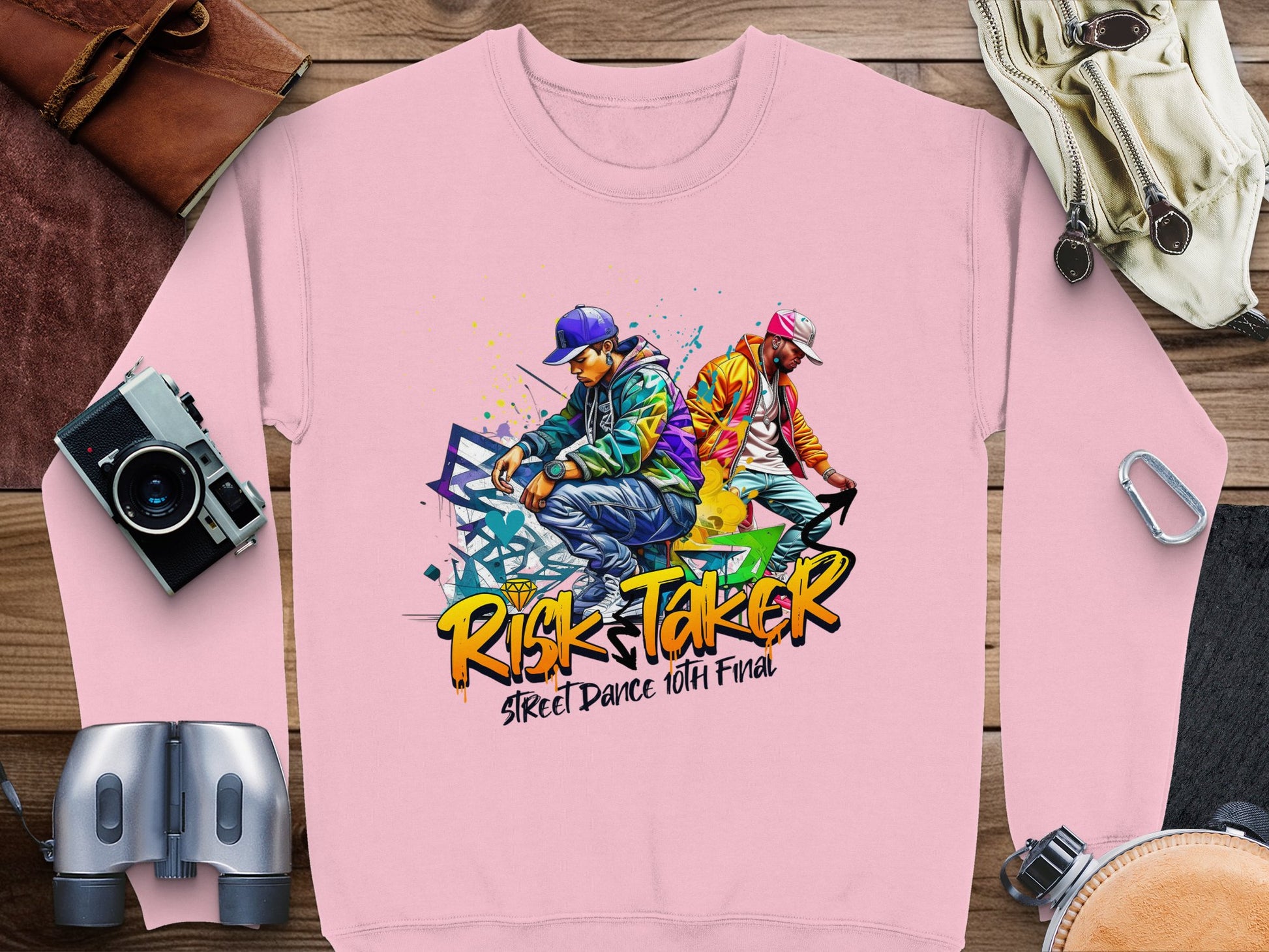 A pink Garment Graphics sweatshirt, featuring two vibrant street dancers and celebrating the Risk Taker Street Dance Finals, is paired with a camera, binoculars, a leather-bound book, bag, and buckle for a unique ensemble.