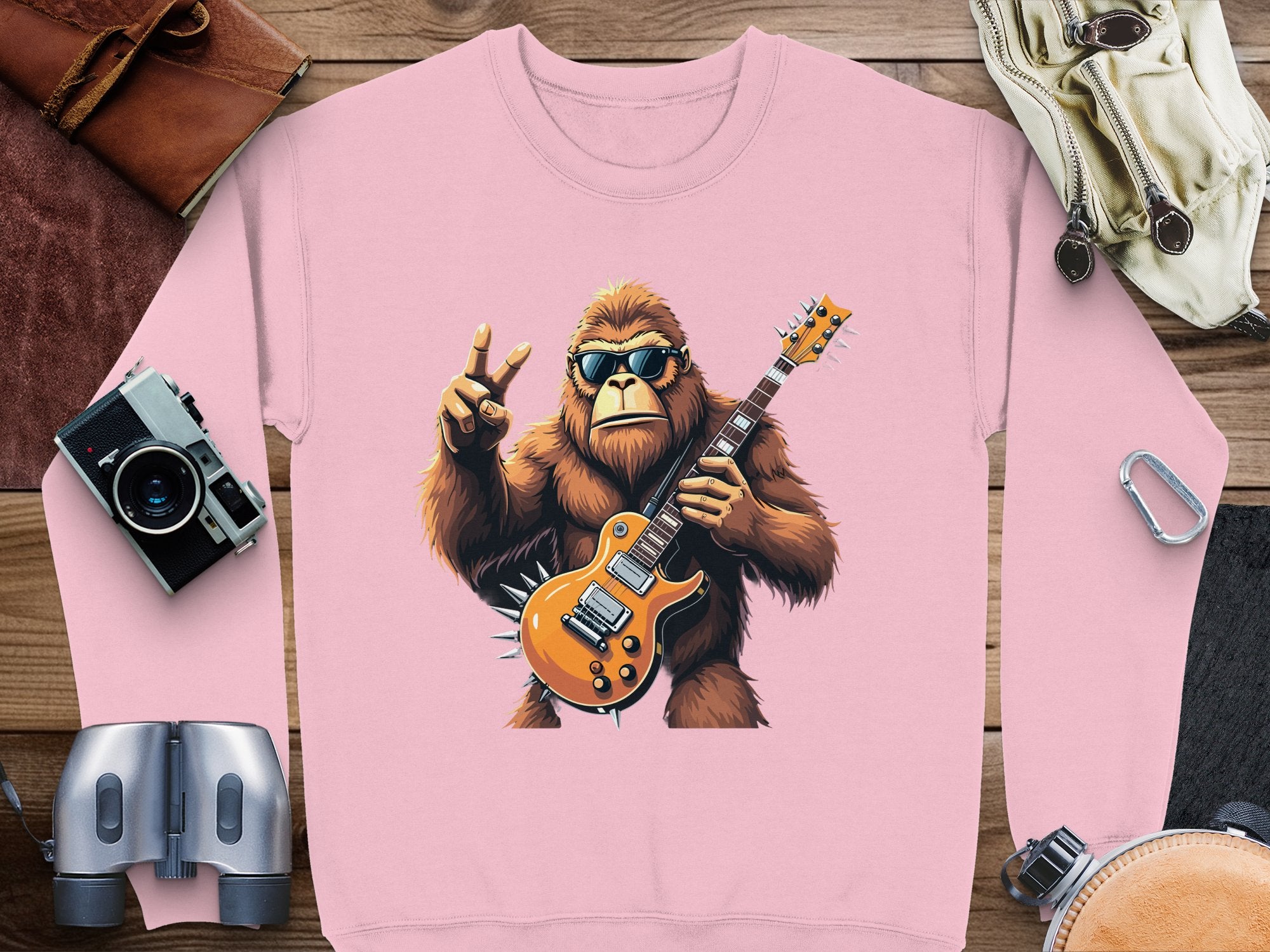 Garment Graphics rocking gorilla sweatshirt showcases a pink design of a gorilla with sunglasses, holding an electric guitar, and flashing a peace sign. Ideal for music lovers, it is displayed alongside a camera, binoculars, goggles, wrench, and leather pouch on a wooden surface.