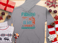 Garment Graphics Anglers Wardrobe gray hoodie features Fishing is my anger management, with a fish and rod graphic, displayed among Christmas decor like gifts, a sweater, pinecones, candy, and ornaments on wooden background.