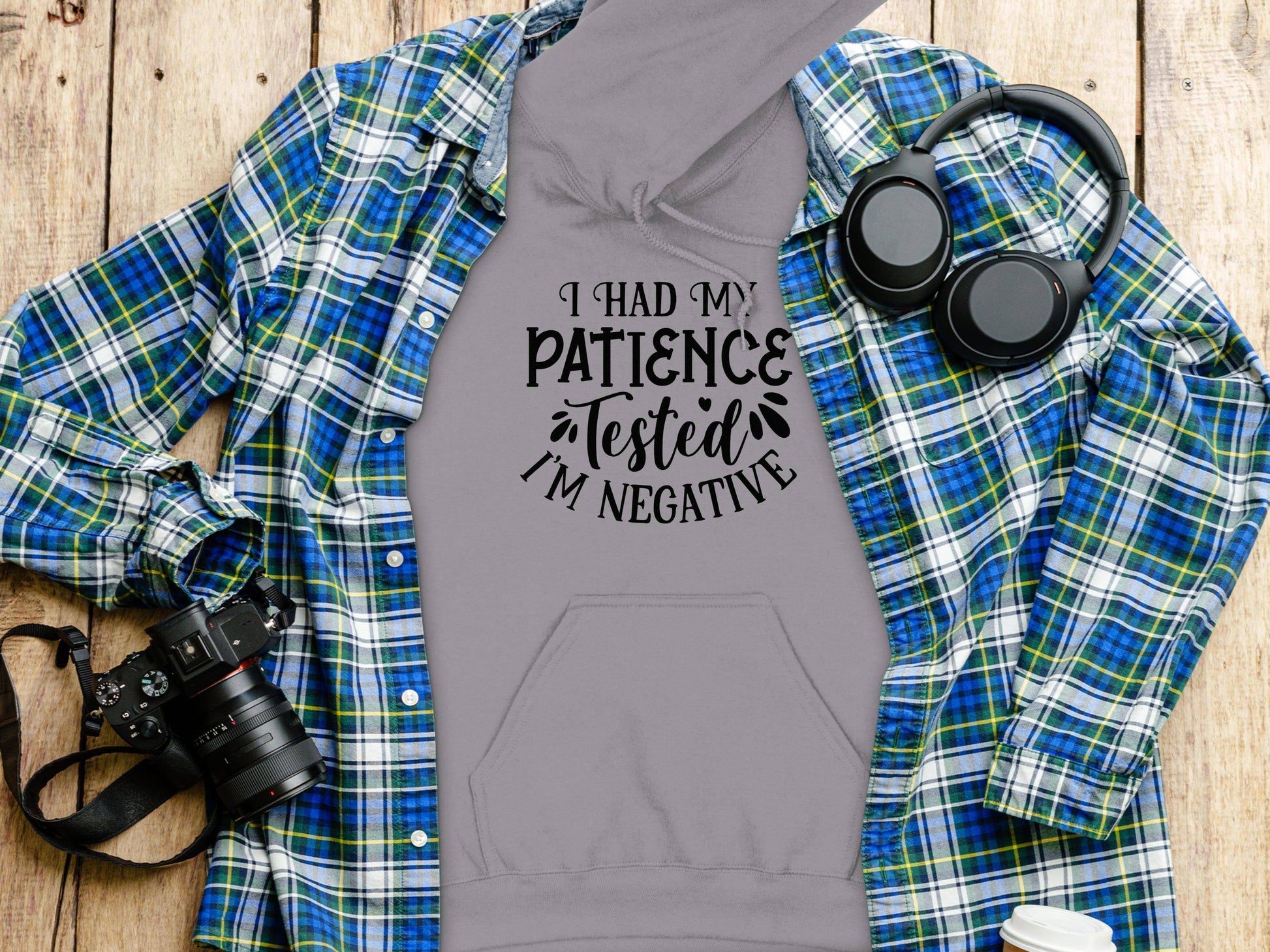 The Garment Graphics Patience Tested Hoodie boasts a classic fit with medium-heavy fabric and the phrase I had my patience tested, Im negative, peeking through a plaid shirt. Nearby are a camera, headphones, and a white cup on a wooden surface.