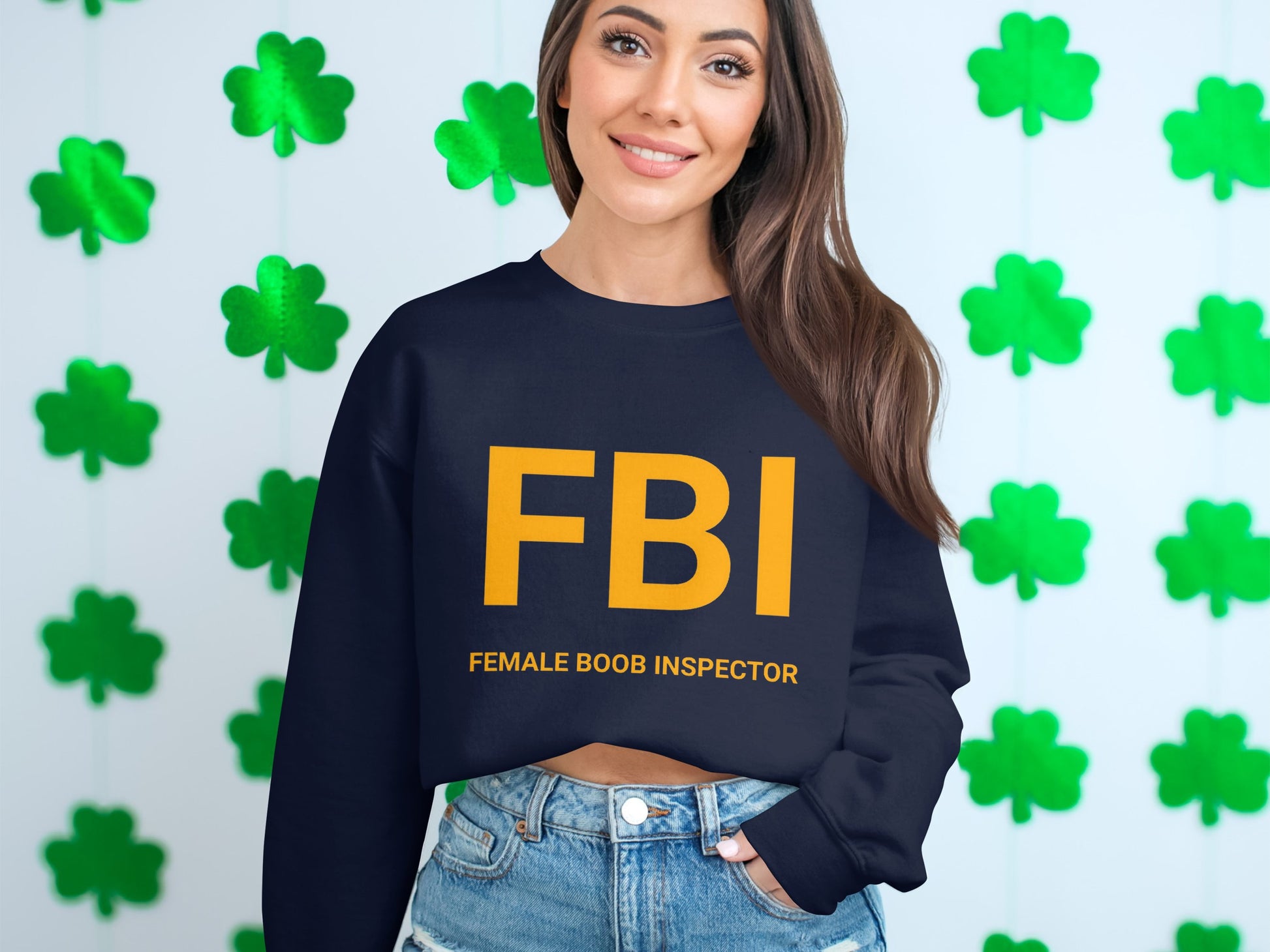 A woman smiles in a Garment Graphics navy blue sweatshirt with FBI Female Boob Inspector emblazoned in bold yellow, set against a playful green shamrock background.