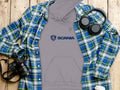 A flat lay features a gray hoodie with the iconic Scania logo by Garment Graphics, paired with a blue and green plaid shirt, black headphones, a black camera, and a white cup lid, all artfully arranged on a wooden surface.