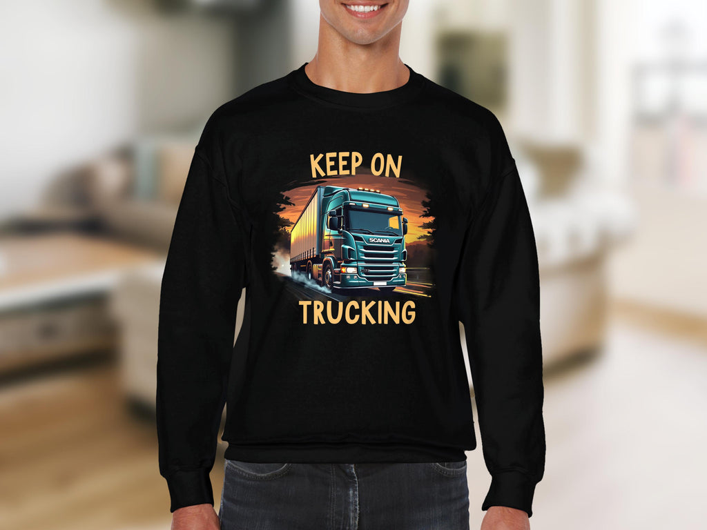 Wearing a Garment Graphics black graphic sweatshirt featuring a bold truck design with KEEP ON TRUCKING over a sunset, they stand indoors radiating casual charm.