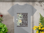 Garment Graphics offers a womens gray t-shirt featuring a cartoon alligator with a basketball and the phrase See you later alligator, set on concrete with greenery, red flowers on the left, and yellow on the right.
