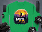 Garment Graphics green retirement sweatshirt features a hiker against a sunset and trees, with Retired 2025 Not My Problem Anymore text. Unisex sizing, displayed with a phone, watch, speaker, shoes, and dumbbells on a gray surface.