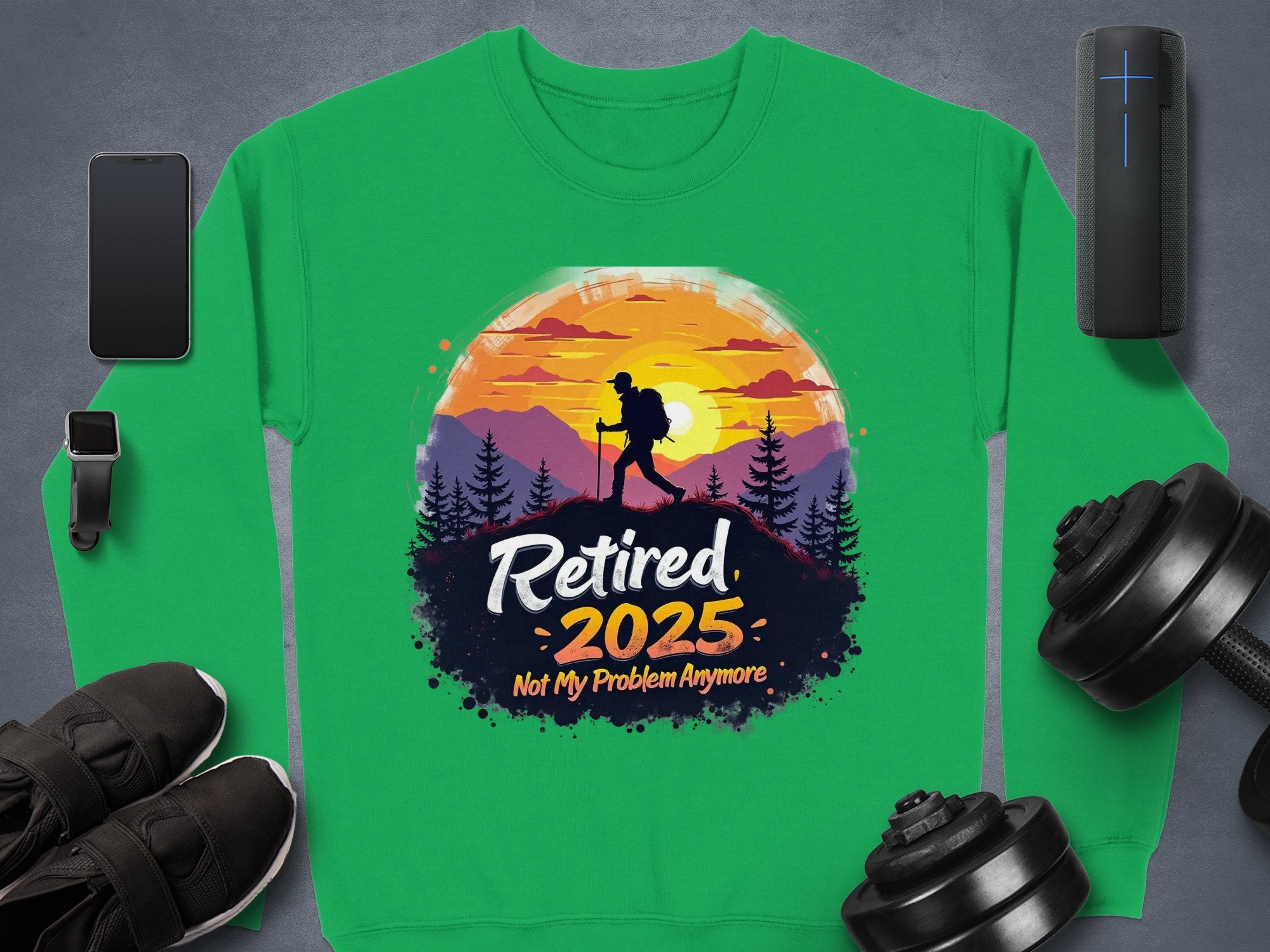 Garment Graphics green retirement sweatshirt features a hiker against a sunset and trees, with Retired 2025 Not My Problem Anymore text. Unisex sizing, displayed with a phone, watch, speaker, shoes, and dumbbells on a gray surface.