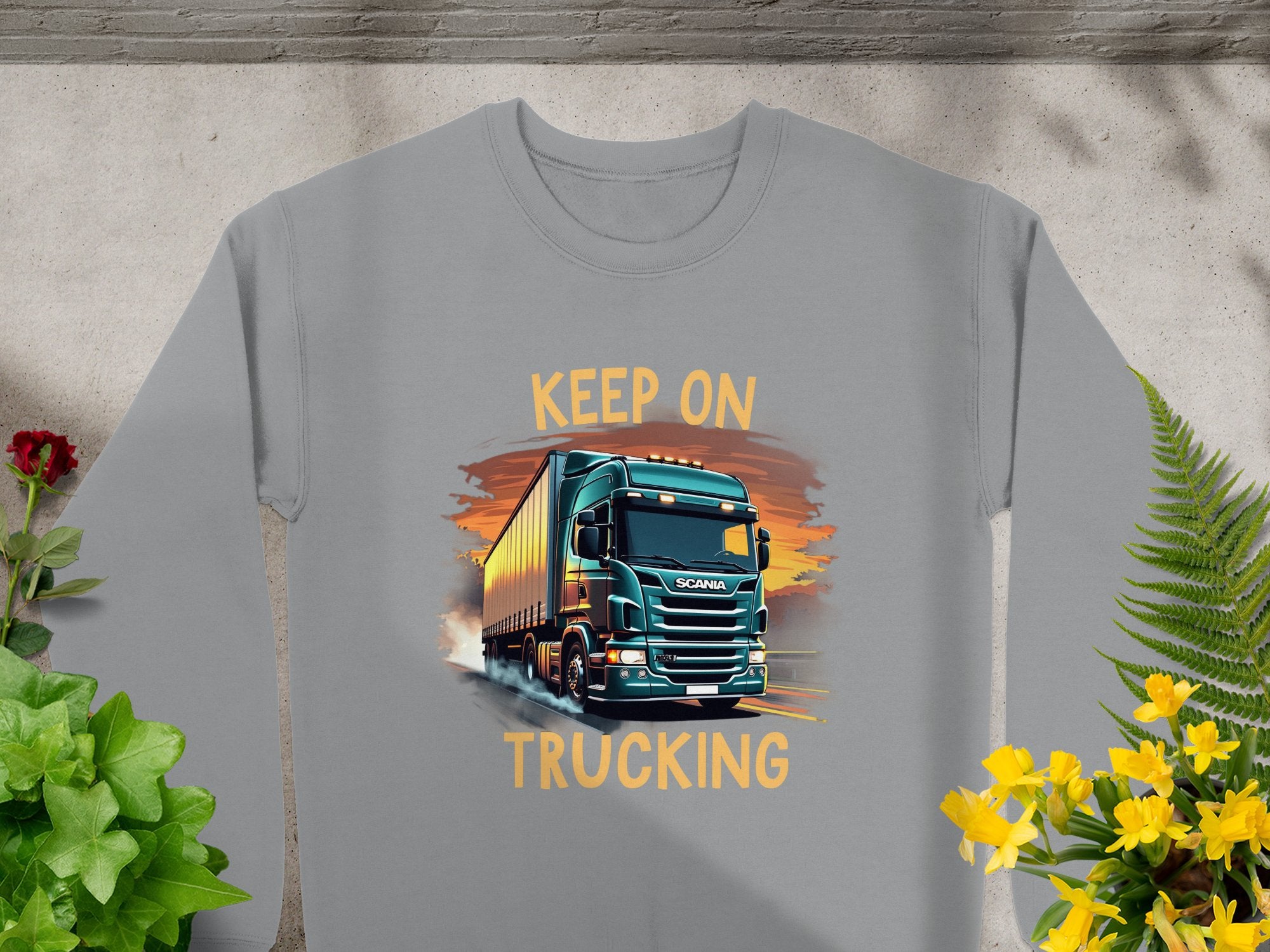 Perfect for truck enthusiasts, Garment Graphics offers a gray sweatshirt featuring a large truck on a road. Adorned with bold orange KEEP ON TRUCKING text, its surrounded by green plants and yellow flowers, making it a unique piece in Graphic Sweatshirts.