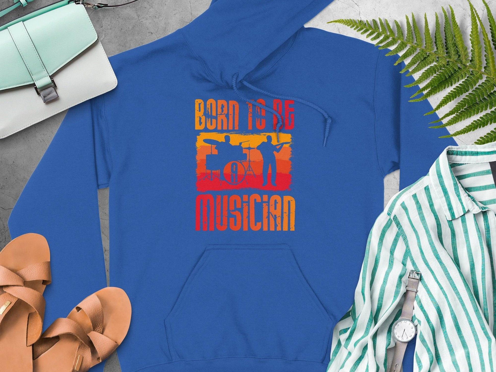 Garment Graphics offers a blue unisex hoodie featuring Born to be a Musician with orange and yellow silhouettes, ideal for music lovers. Pair it effortlessly with a mint bag, brown sandals, striped shirt, fern leaf accessory, watch, and glasses on a gray surface.